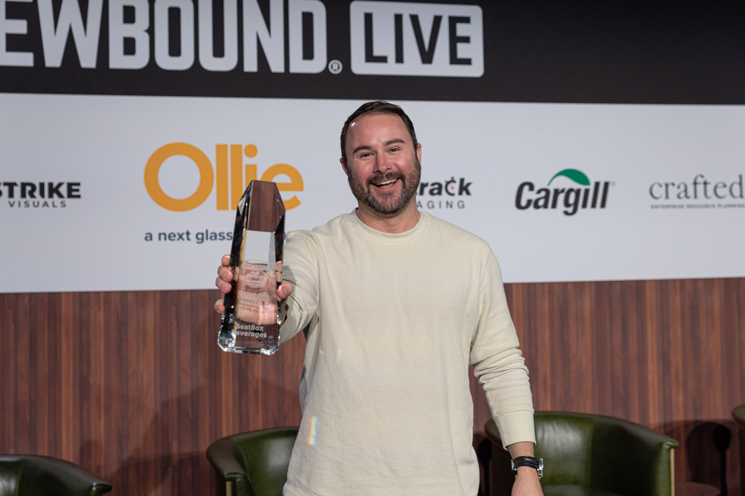 A smiling man in a cream sweater holds up a glass award in front of the Brewbound Live sponsor backdrop.