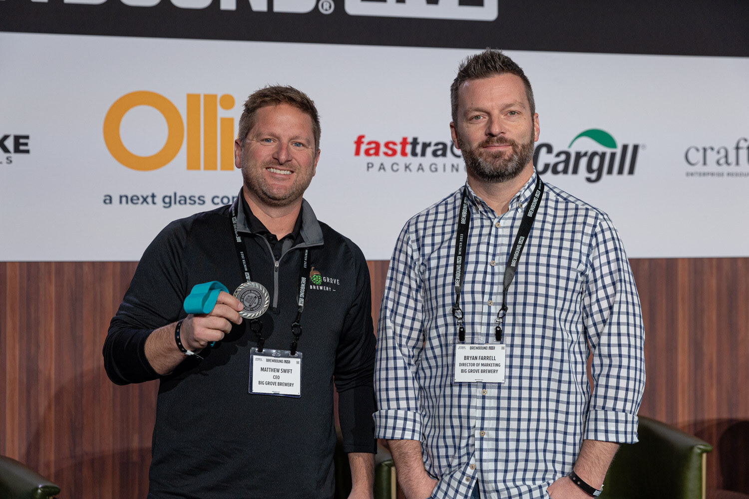 Two men wearing event lanyards stand side by side, one holding a silver medal, in front of the Brewbound Live backdrop.