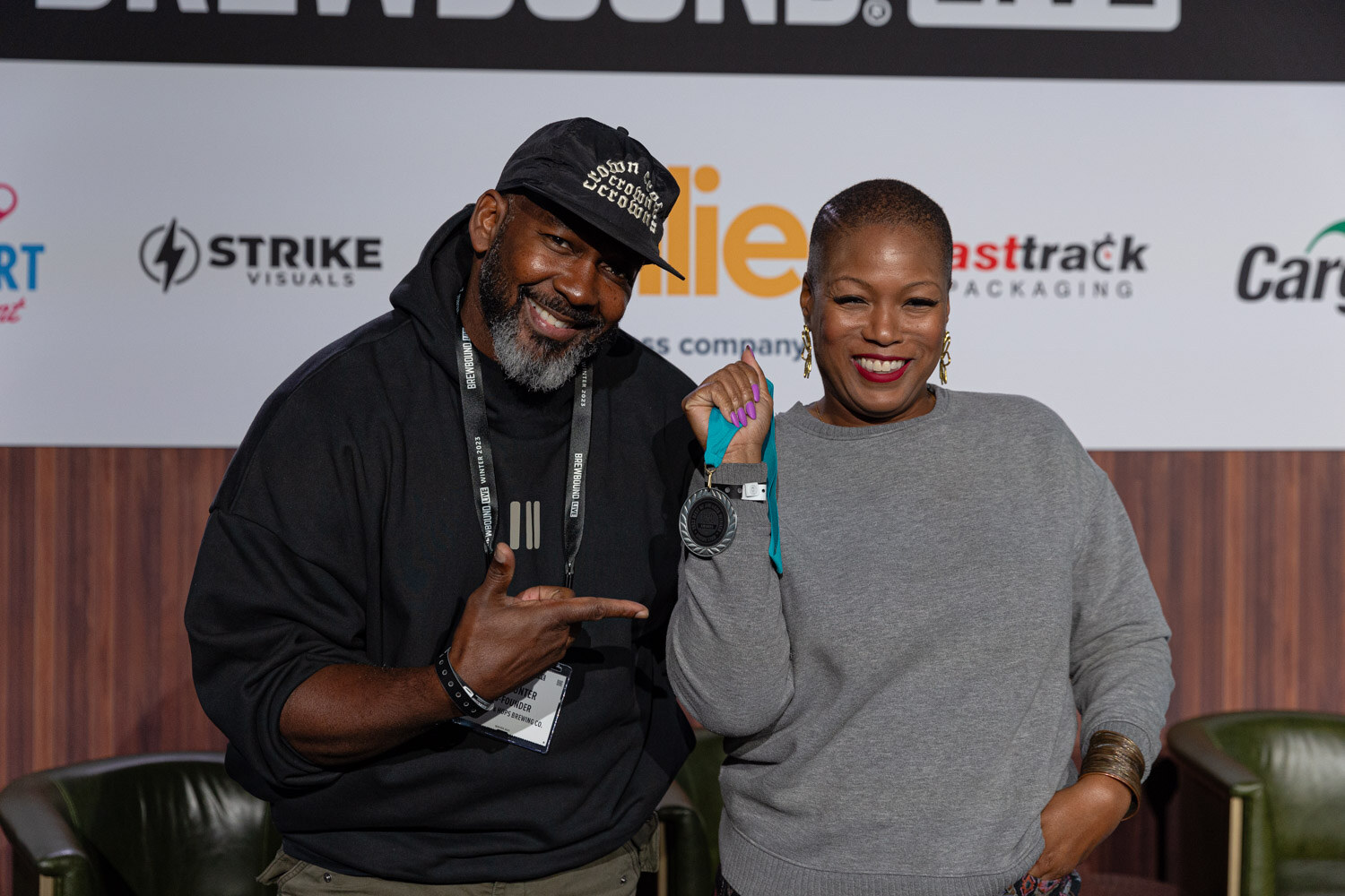 A smiling man in a black hoodie and cap and a woman in a gray sweater pose together on stage.