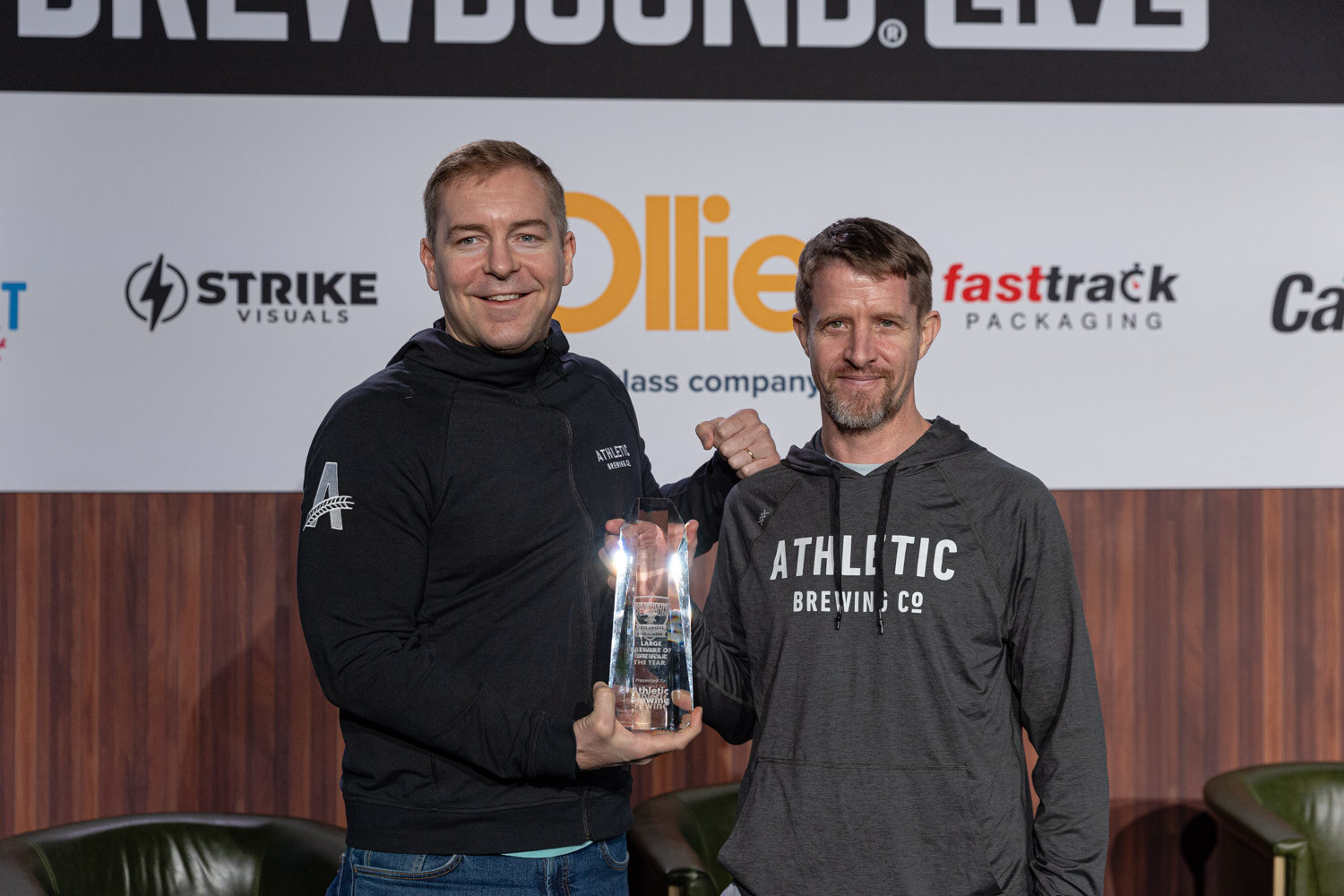 Two men in black Athletic Brewing hooded tops pose with a glass award on stage in front of the Brewbound Live backdrop.