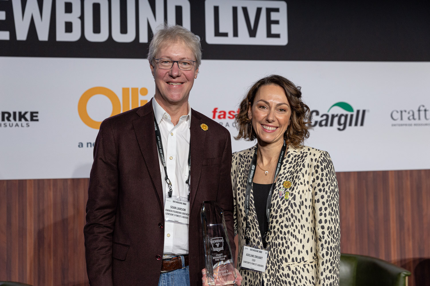 A man in a burgundy blazer and a woman in a leopard-print jacket smile with a glass award in front of the Brewbound Live backdrop.