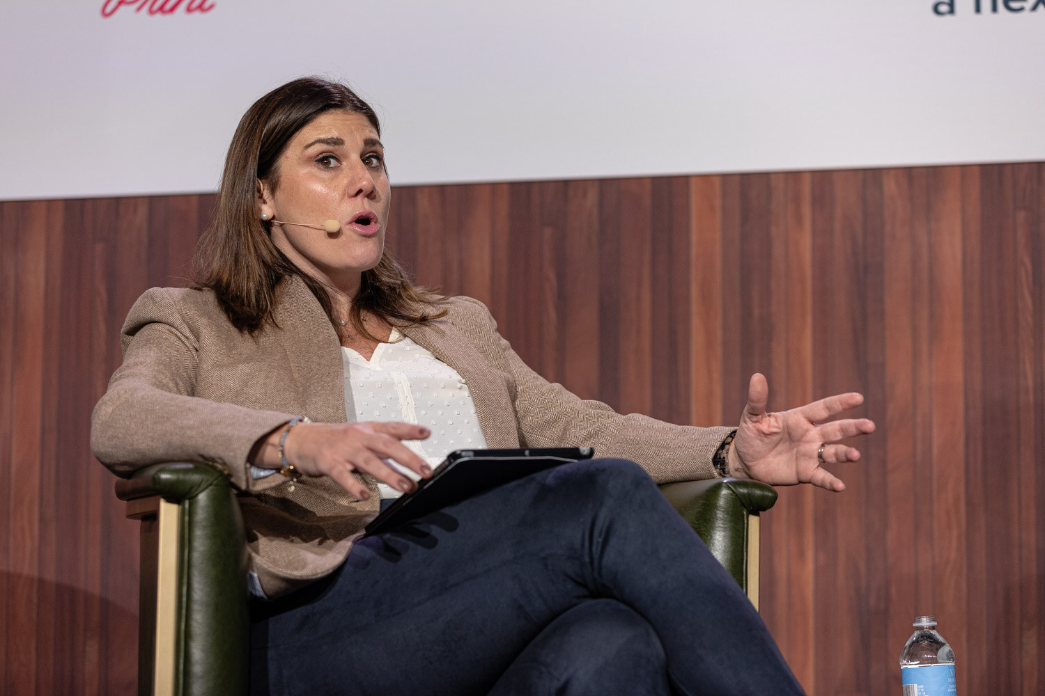 A woman in a tan blazer sits in a green armchair holding a tablet and gesturing with both hands.