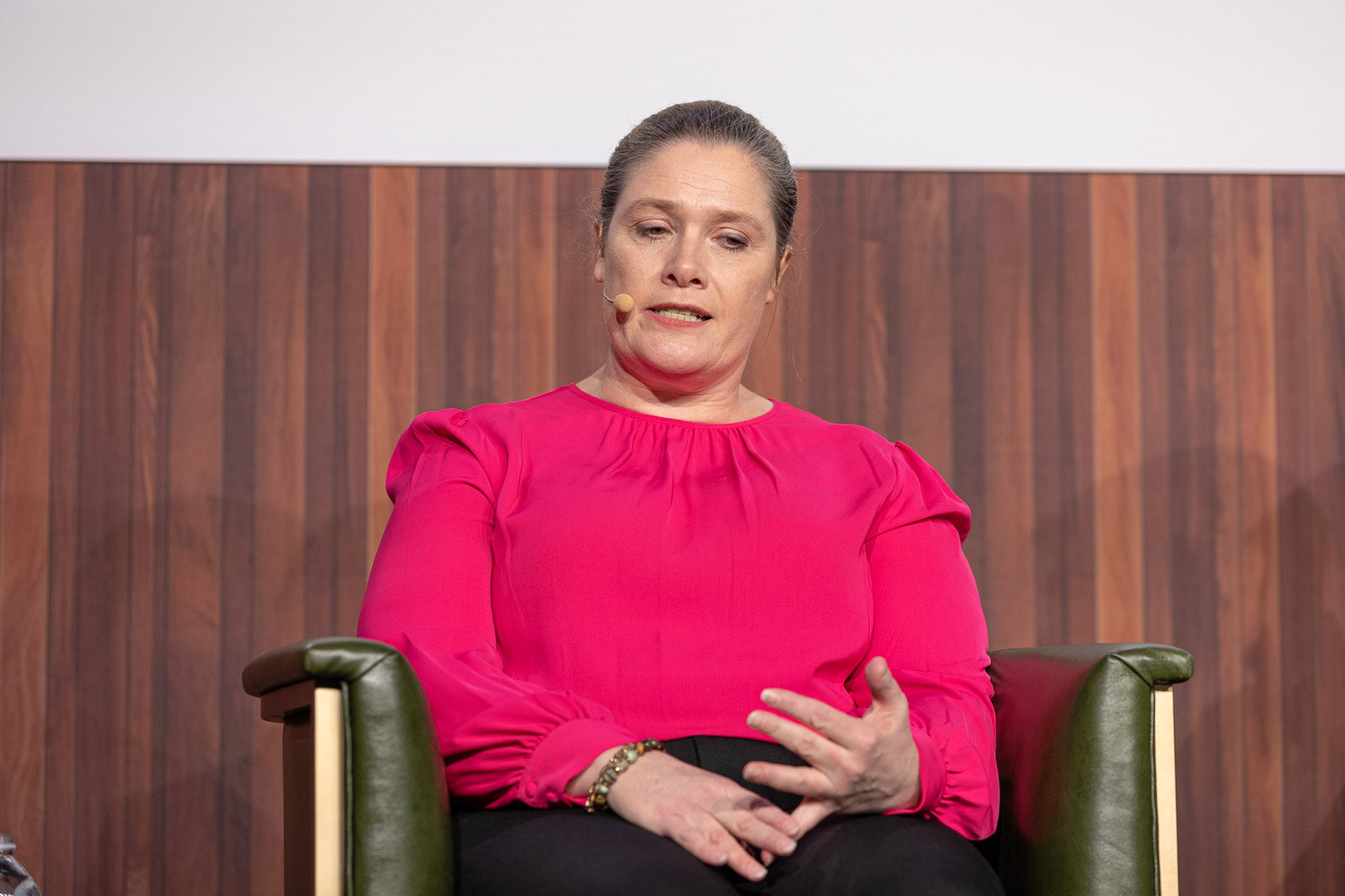 A woman in a pink blouse sits with her hands together in a green armchair against a wood-paneled stage wall.