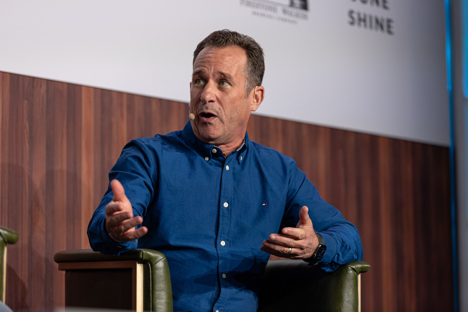 A man in a blue button-up shirt gestures with both hands while seated in an armchair on stage.