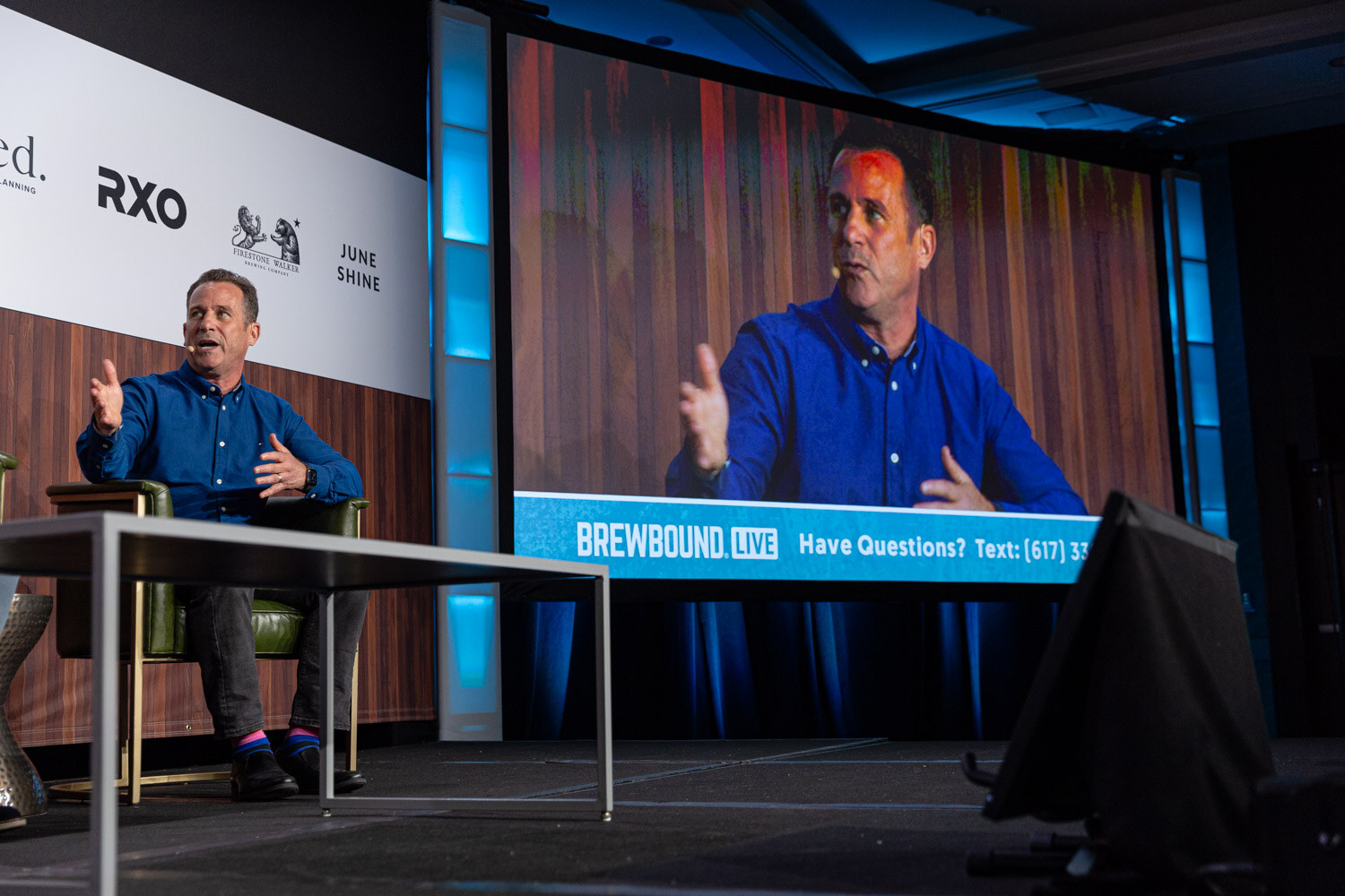 A man in a blue shirt sits on stage while a large screen beside him shows a close-up of the same speaker with a Have Questions text banner.