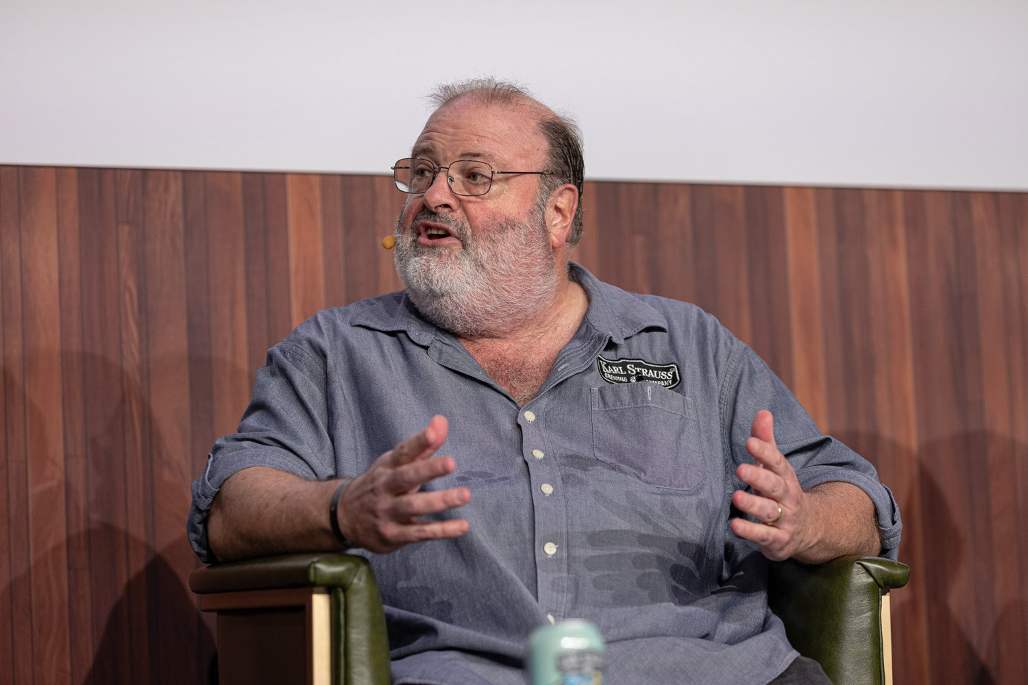 A bearded man in glasses and a gray work shirt gestures while speaking in an armchair.