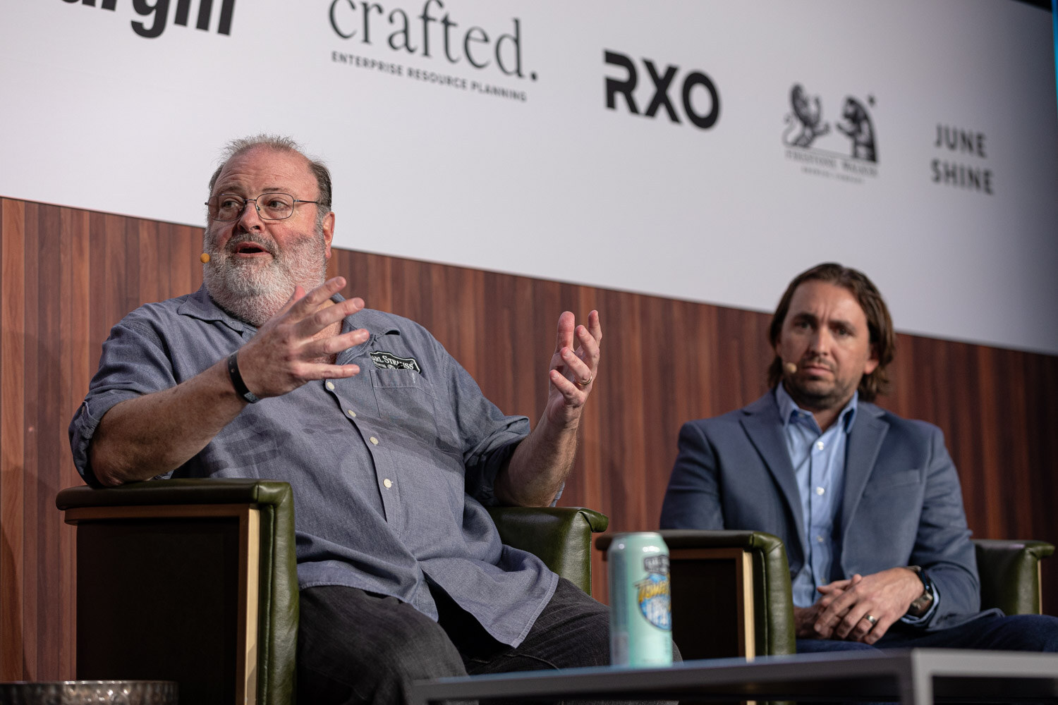 A bearded man in glasses gestures while a man in a blue blazer sits beside him, with a beer can on a table in front.