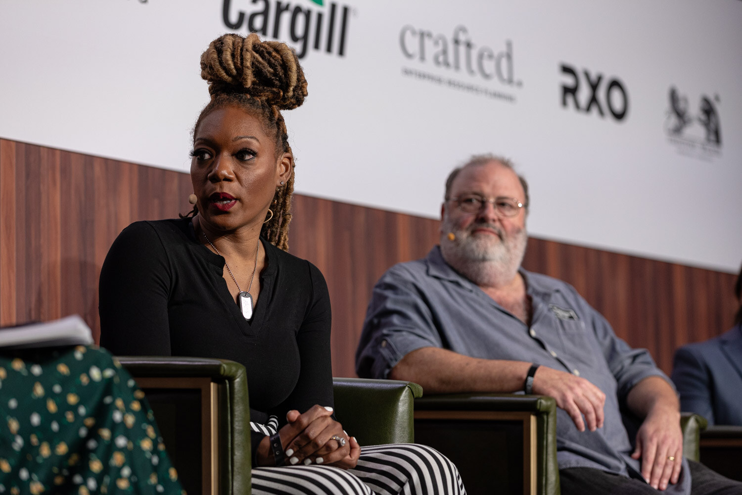 A woman in a black top with braided hair and a bearded man in glasses sit in armchairs on stage.