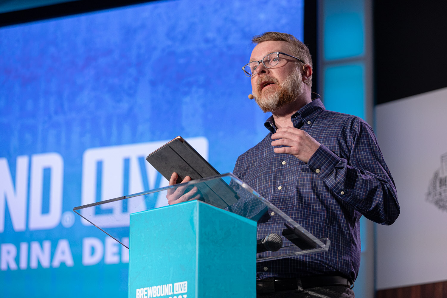 A bearded man in glasses and a plaid shirt speaks at a teal lectern holding a tablet in front of a blue screen.