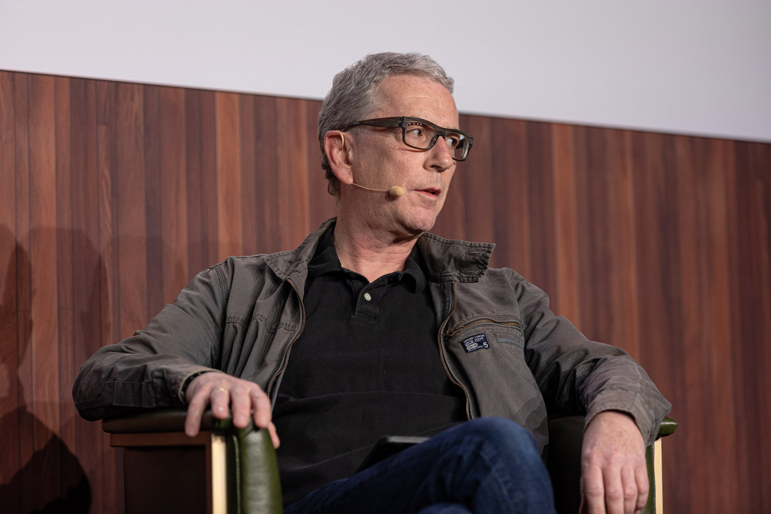 A gray-haired man in glasses and an olive jacket sits in an armchair, looking off to the side.