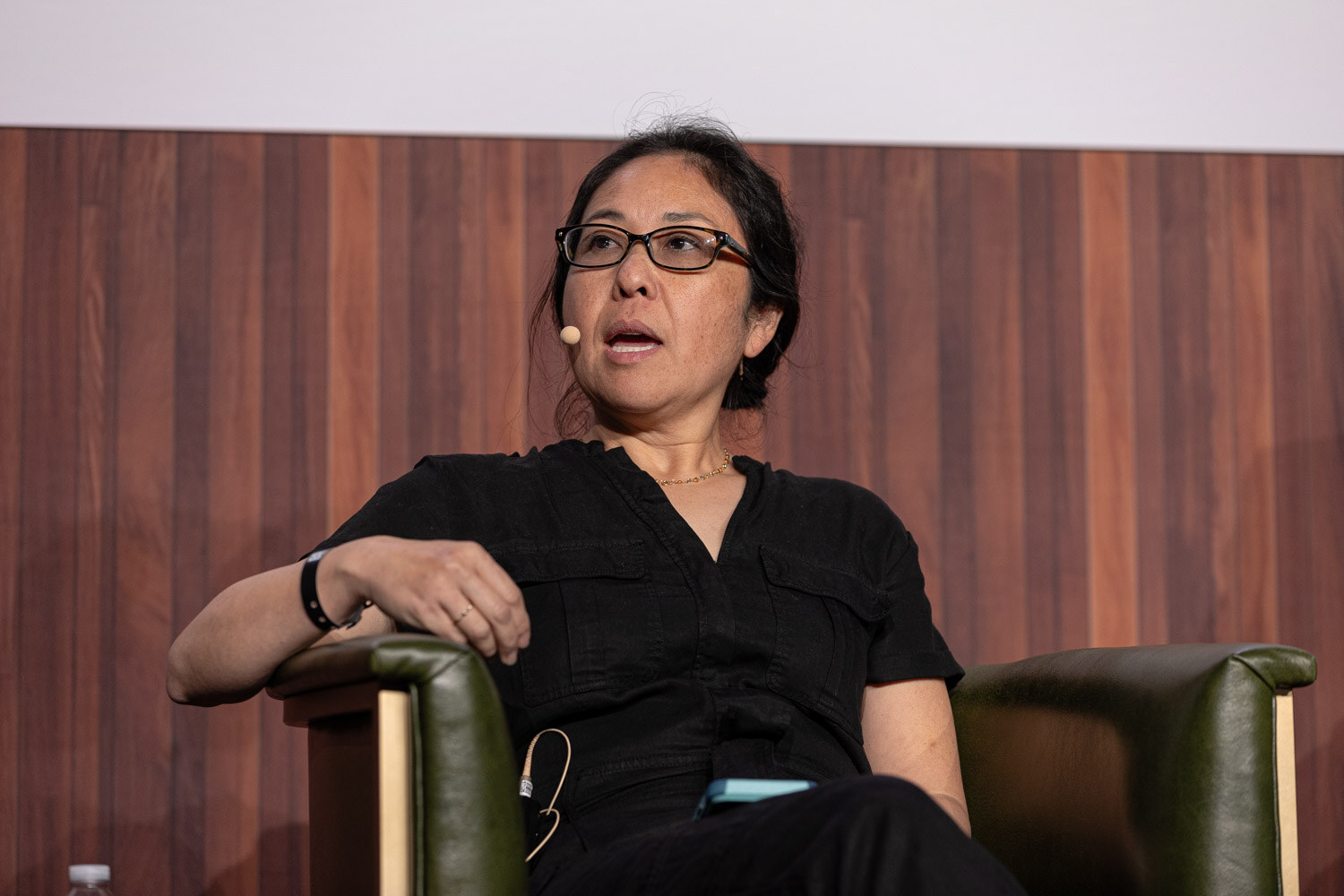 A woman with glasses and a headset microphone sits in an armchair and speaks, wearing a black top.