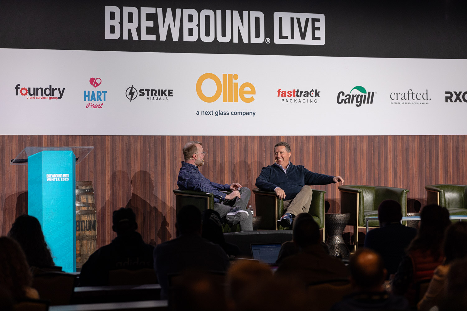 Two men sit in armchairs on stage beneath the Brewbound Live banner with audience heads in silhouette in the foreground.