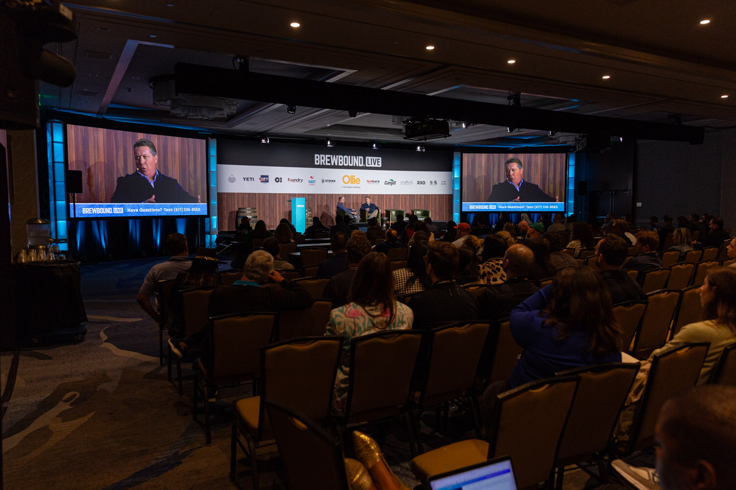 A dim ballroom with rows of seated attendees facing a distant stage and large side screens under the Brewbound Live banner.