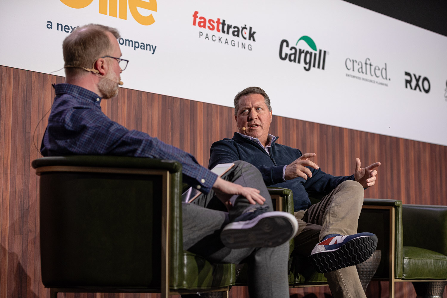 Two men sit in armchairs on stage in conversation, one in a blue plaid shirt and one in a navy quarter-zip.