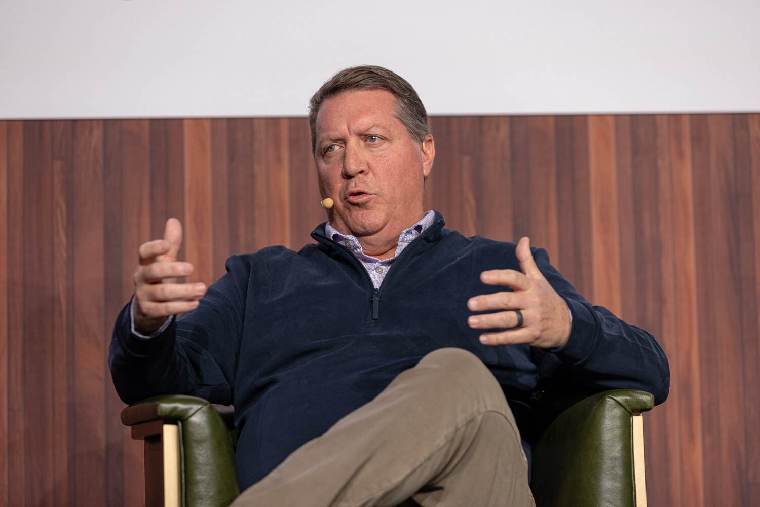 A man in a navy quarter-zip and khakis sits in an armchair with his hands open as he speaks.