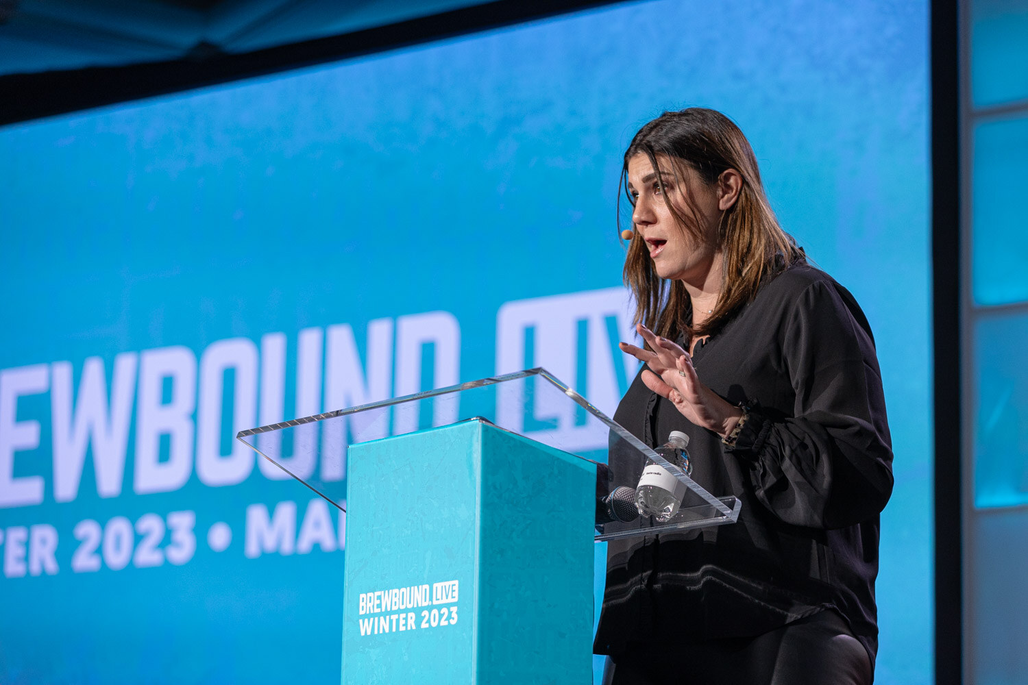 A woman in black speaks at a clear lectern in front of a blue Brewbound Live Winter 2023 screen.