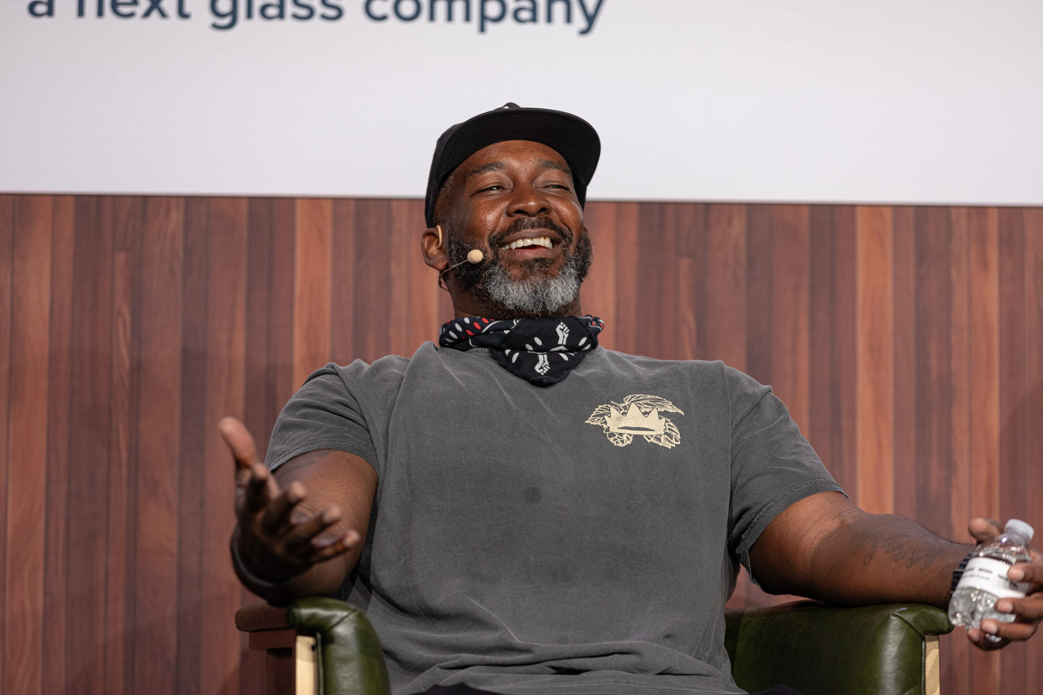 A smiling bearded man in a cap and gray t-shirt holds his arms out wide while seated in an armchair.