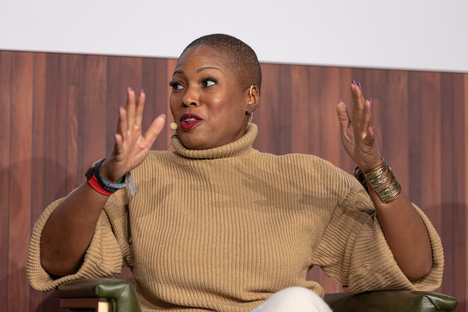A woman with a short shaved hairstyle in a tan turtleneck sweater gestures with both hands while seated.