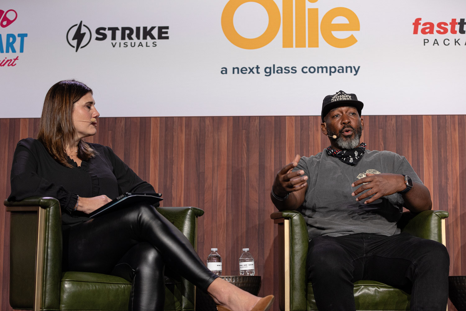 A woman in black holding a tablet sits facing a man in a cap and gray t-shirt in two green armchairs.