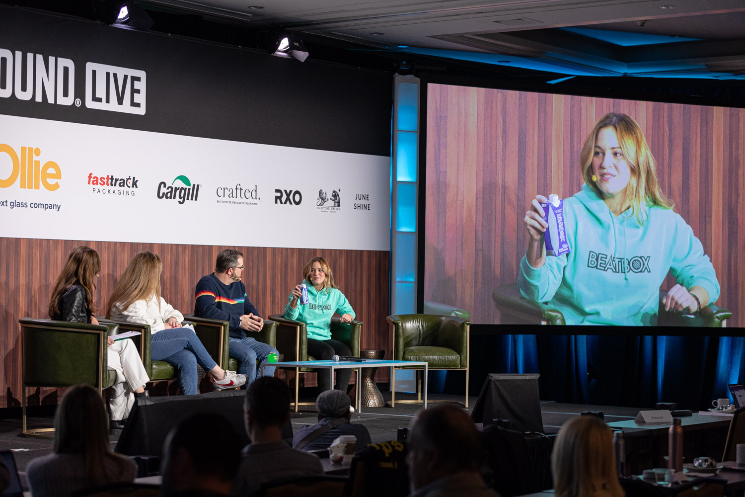 Four people sit on stage in armchairs while a large screen shows a woman in a teal BeatBox hoodie holding a can.