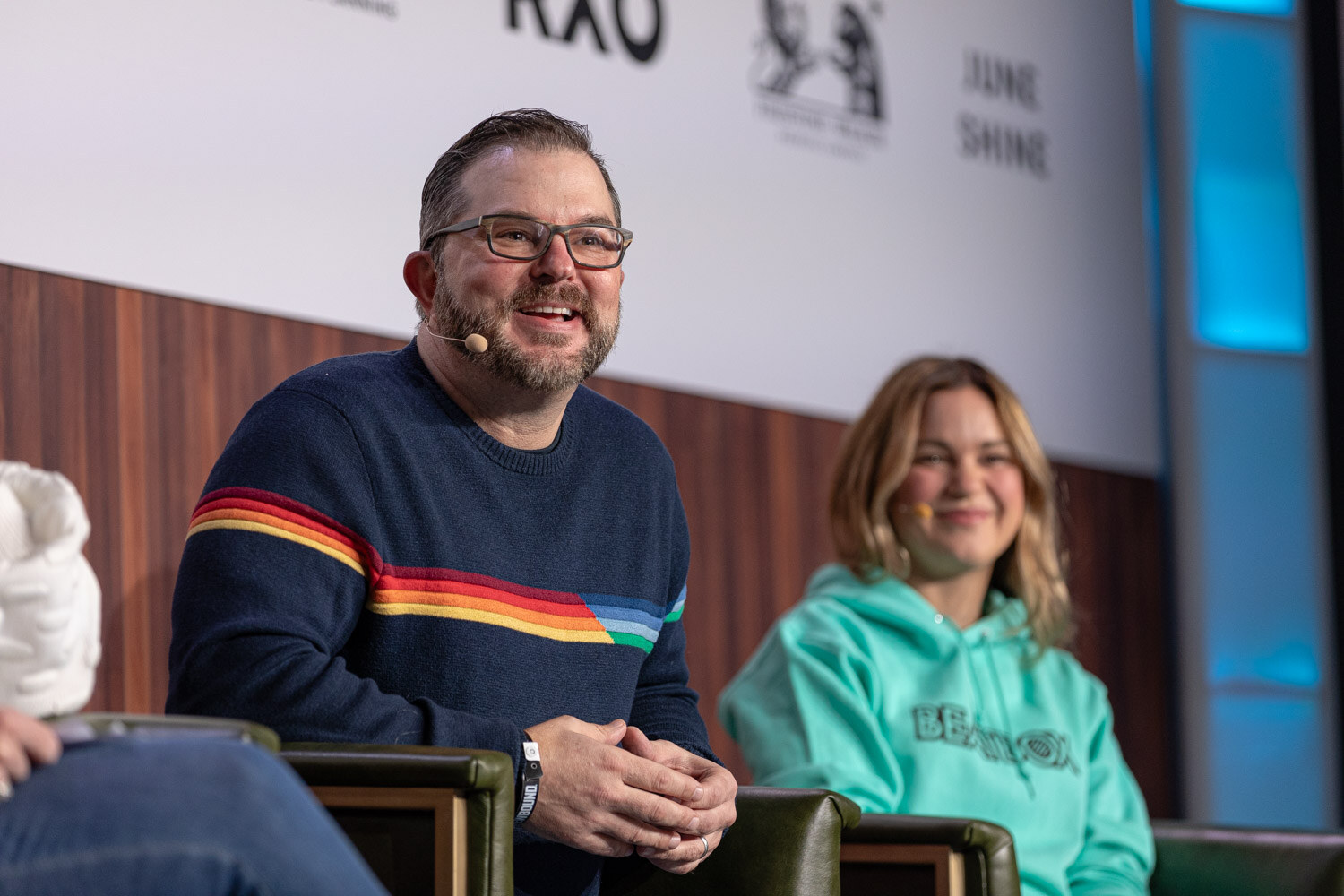 A bearded man in a rainbow-striped navy sweater and a woman in a teal BeatBox hoodie smile on stage.