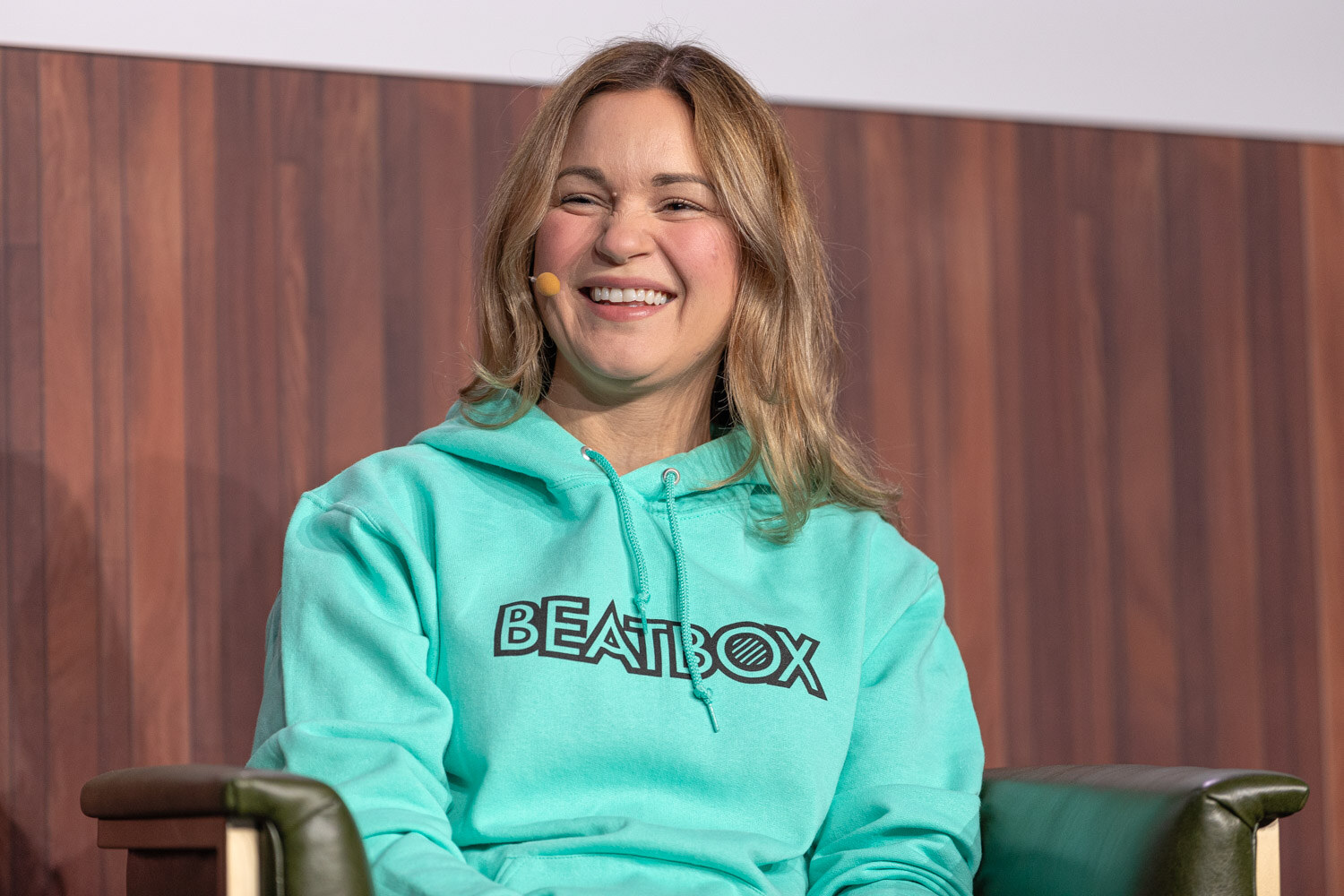 A smiling woman with long blonde hair wears a teal hoodie with the BeatBox logo and sits in a green armchair.