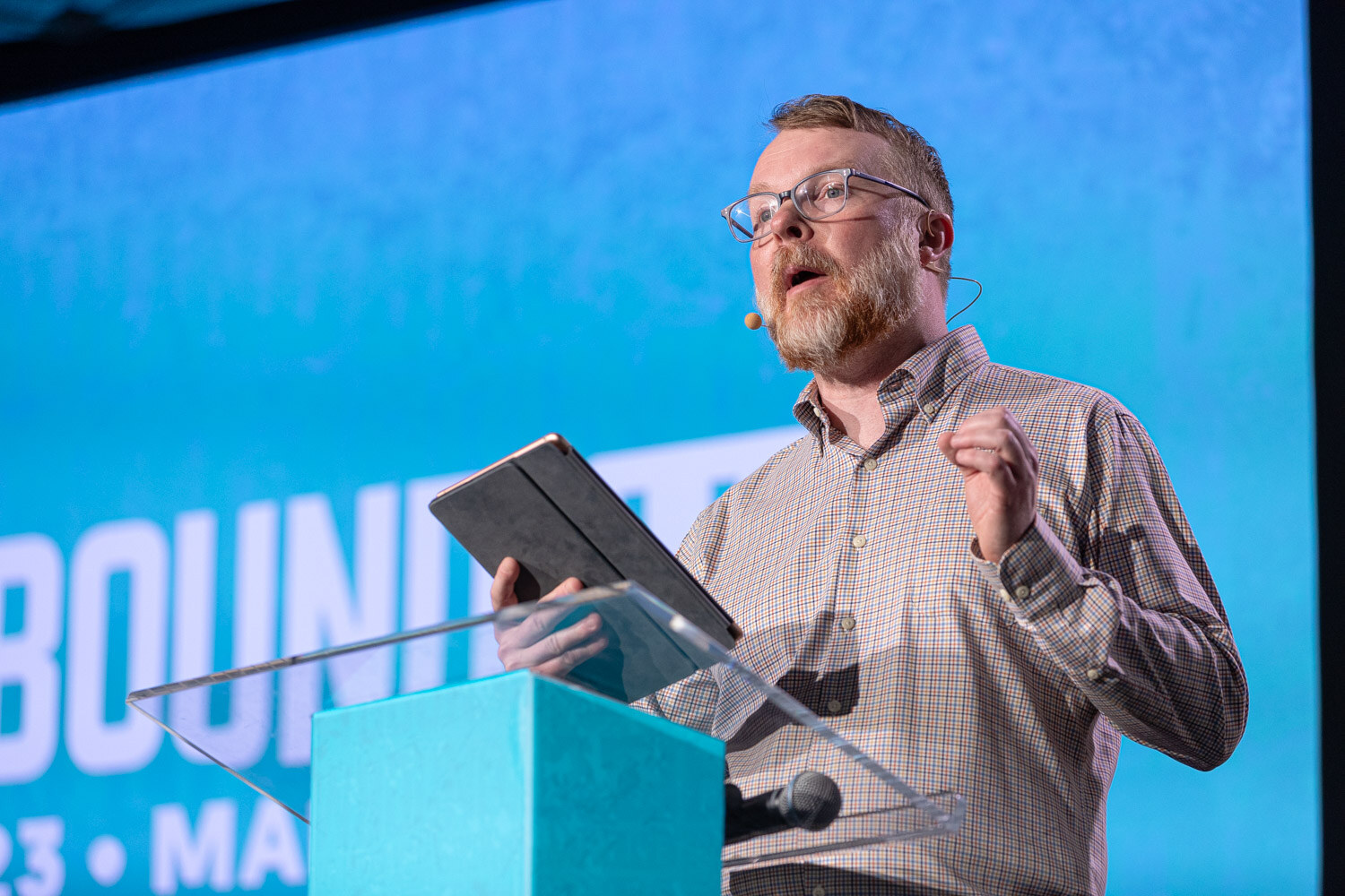 A bearded man in glasses holds a tablet and gestures at a clear lectern in front of a blue Brewbound Live screen.