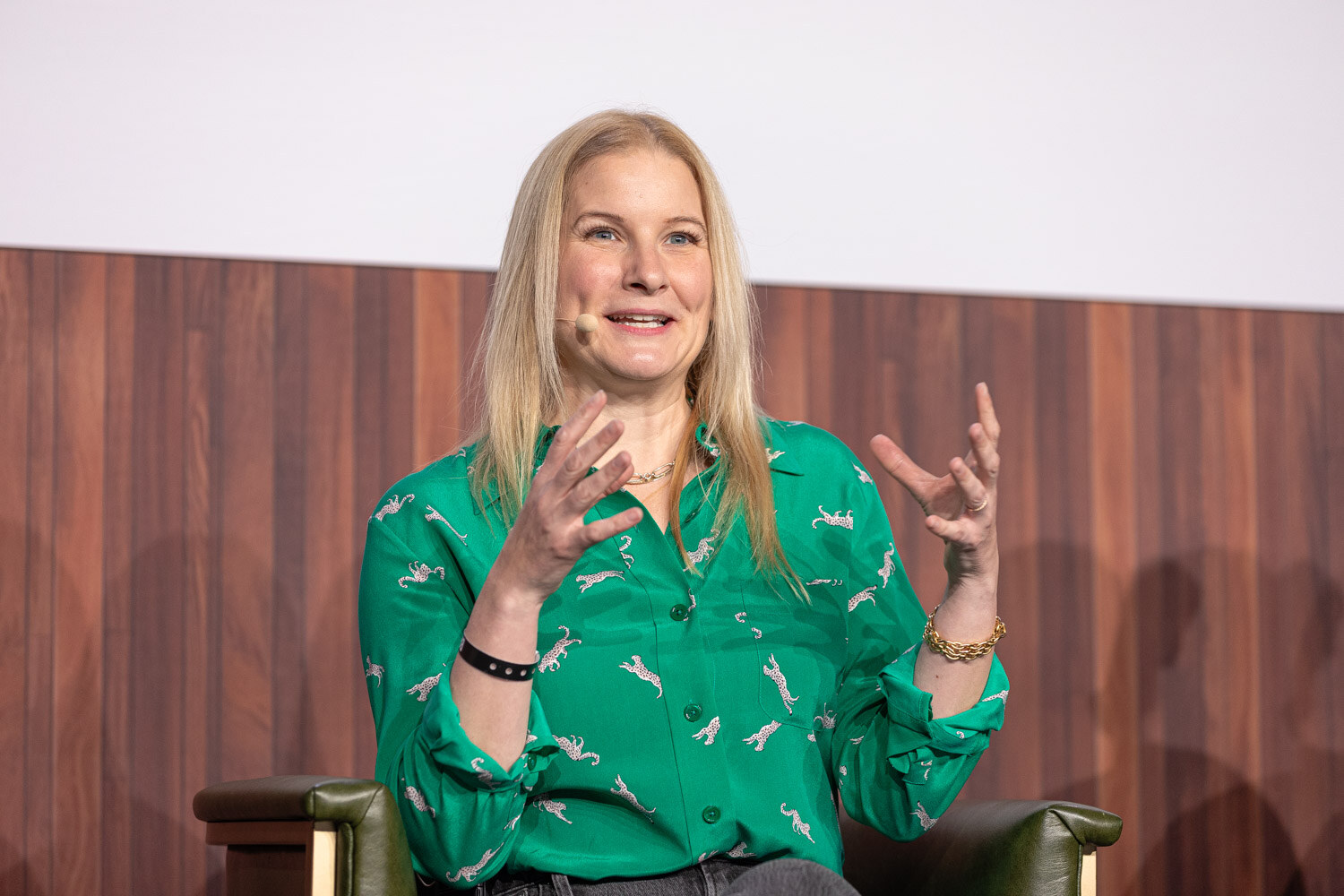 A woman with long blonde hair in a green patterned blouse gestures with both hands while seated.