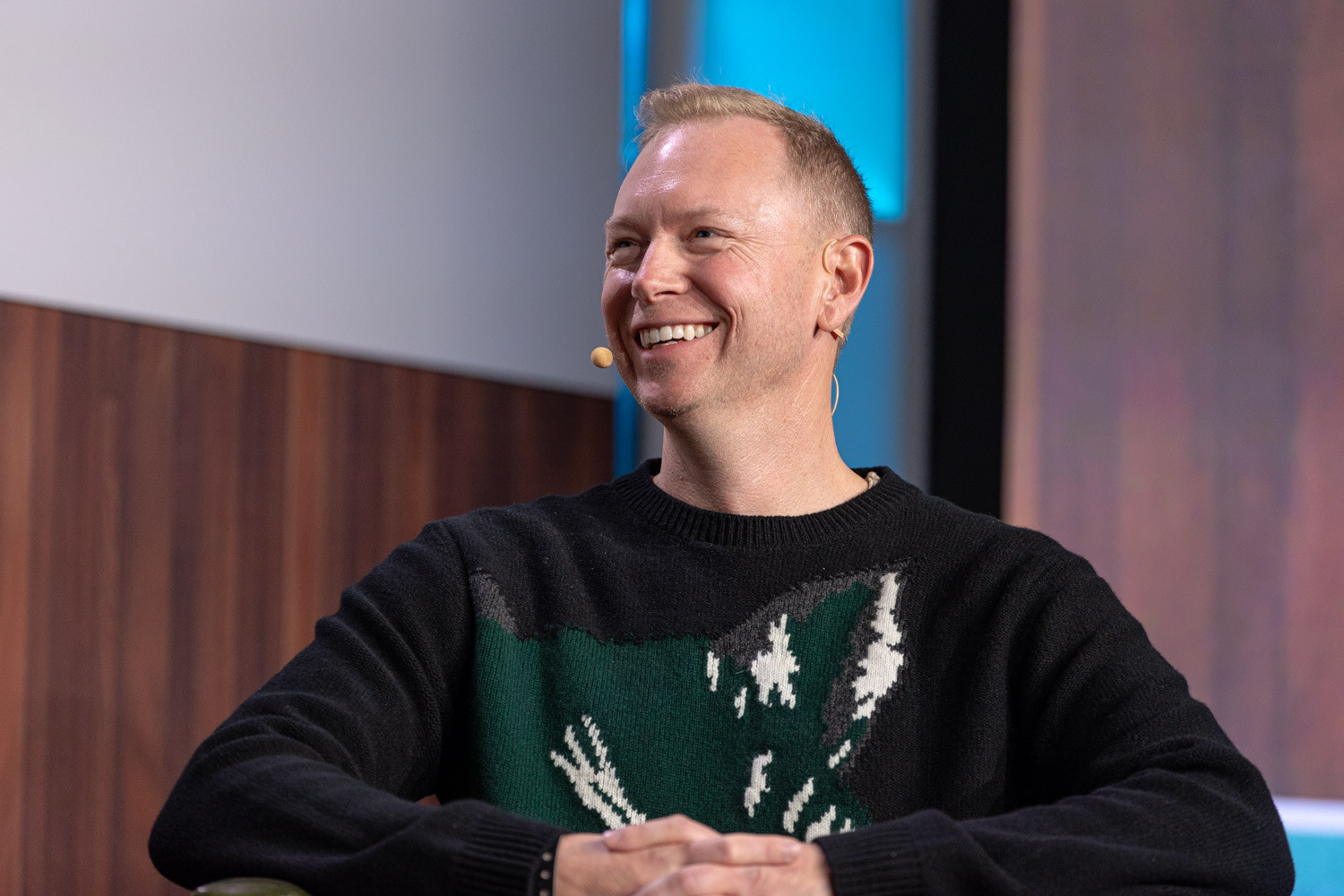 A smiling man in a black sweater with a green animal graphic sits looking off to the side.