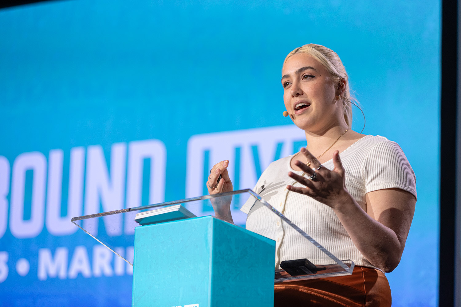 A woman with blonde hair in a white top gestures at a clear lectern in front of a blue Brewbound Live screen.