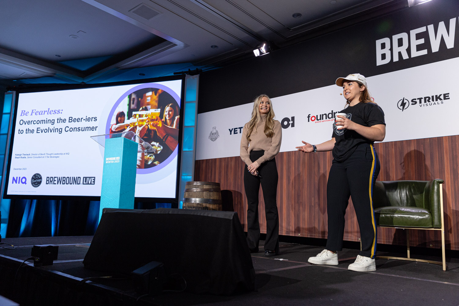 Two women stand on a stage beside a lectern and a screen reading Be Fearless: Overcoming the Beer-iers to the Evolving Consumer.