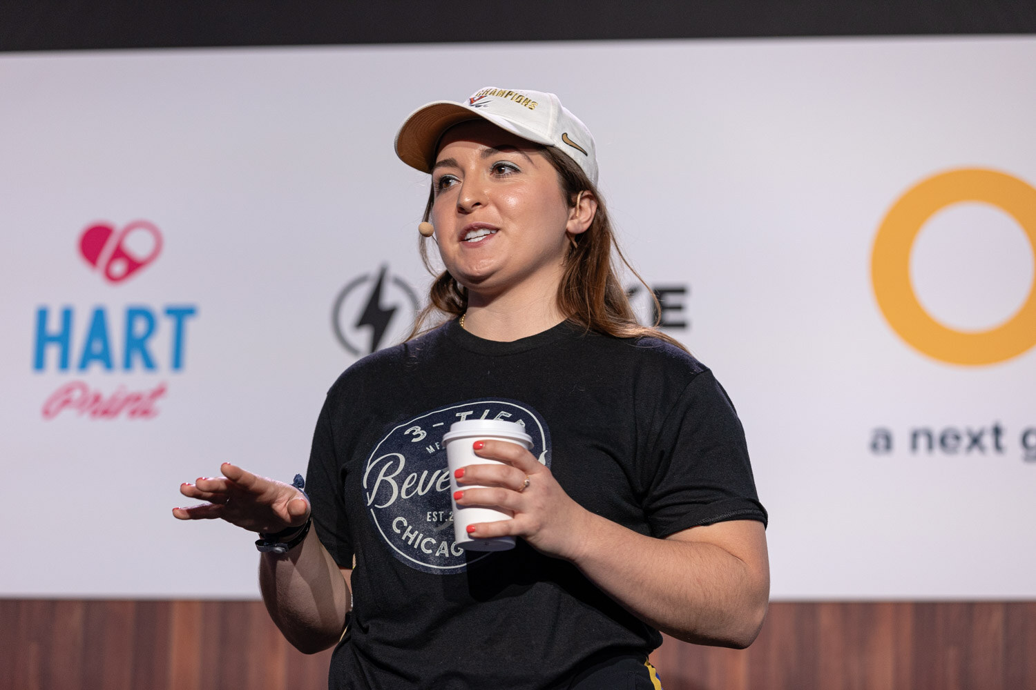 A woman in a baseball cap and black t-shirt holds a paper coffee cup and gestures while speaking.