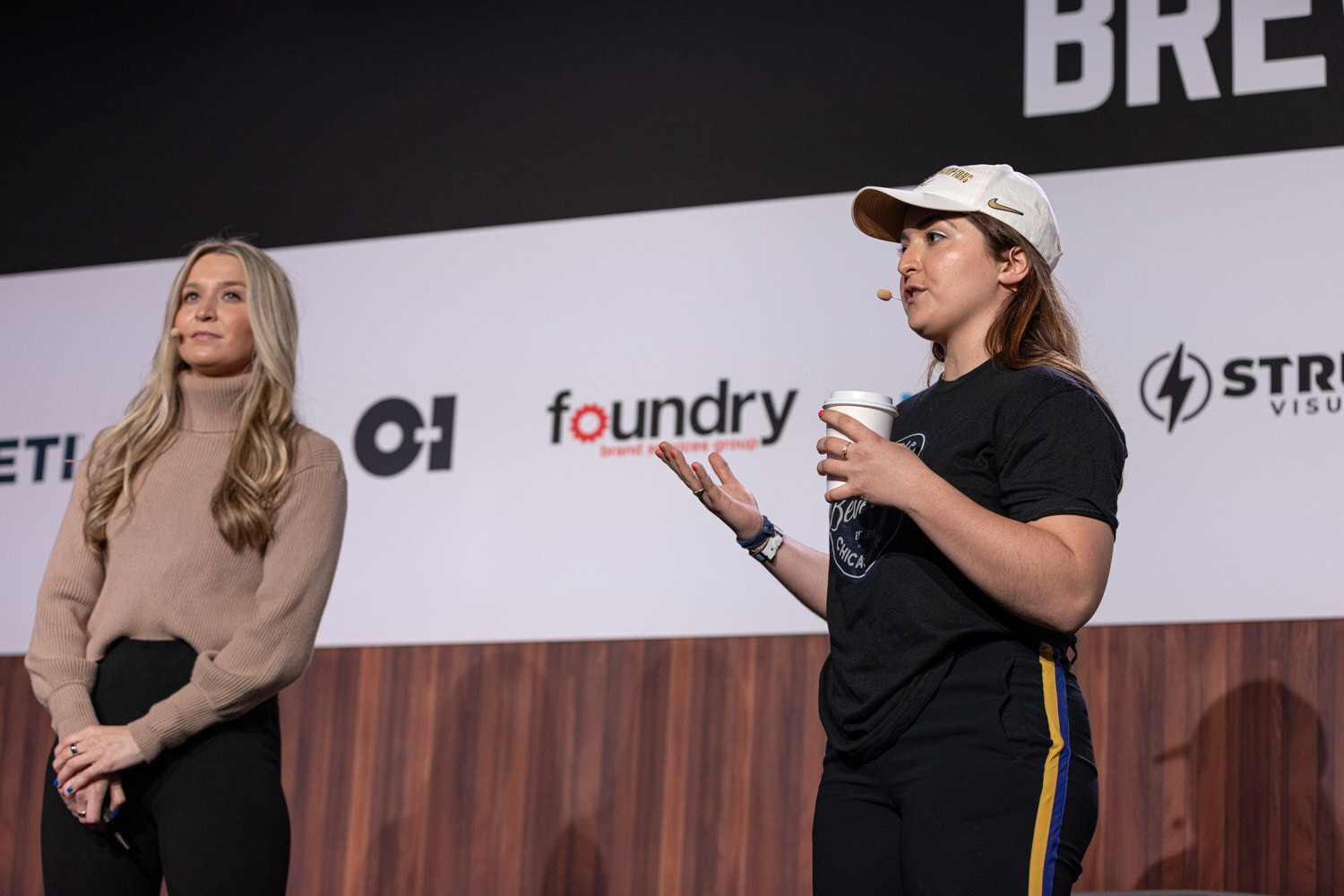 A woman in a tan sweater stands next to a woman in a cap and black t-shirt who speaks while holding a coffee cup.