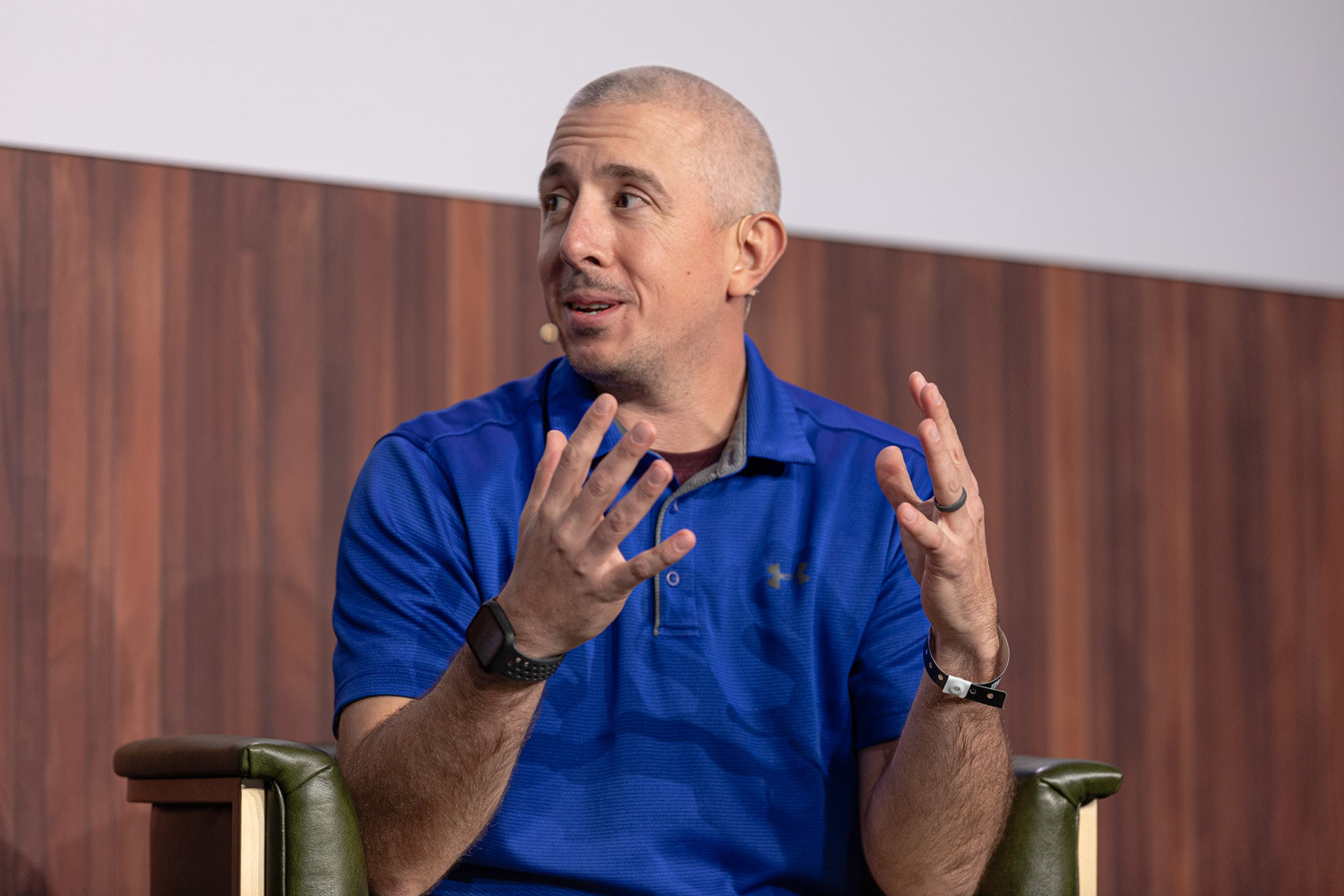 A man in a royal blue polo shirt sits in an armchair and gestures with both hands.