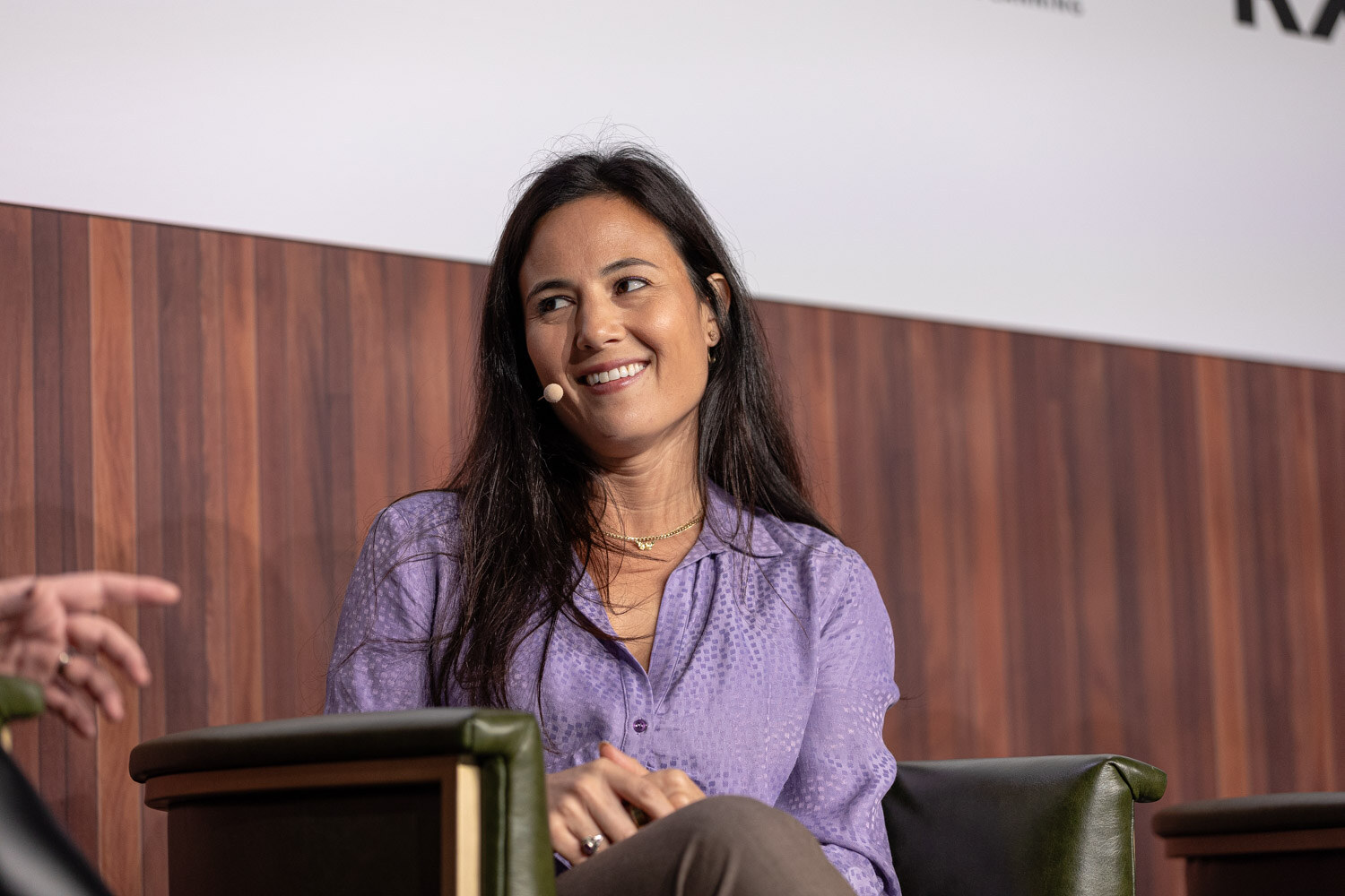 A woman with long dark hair in a lavender blouse smiles while seated in an armchair.