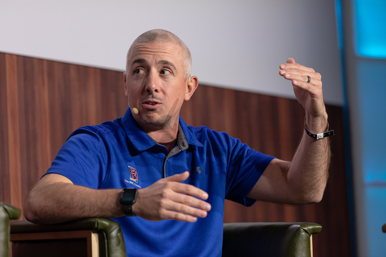 A man in a blue polo with one arm raised gestures while seated in a green armchair.