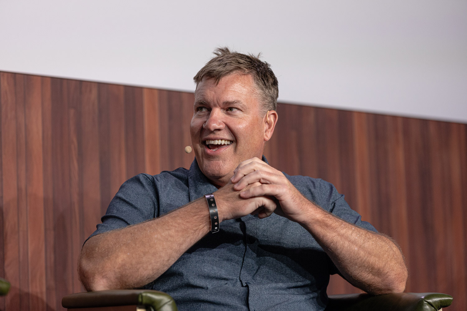 A man in a blue-gray polo shirt smiles with his hands clasped while seated on stage.