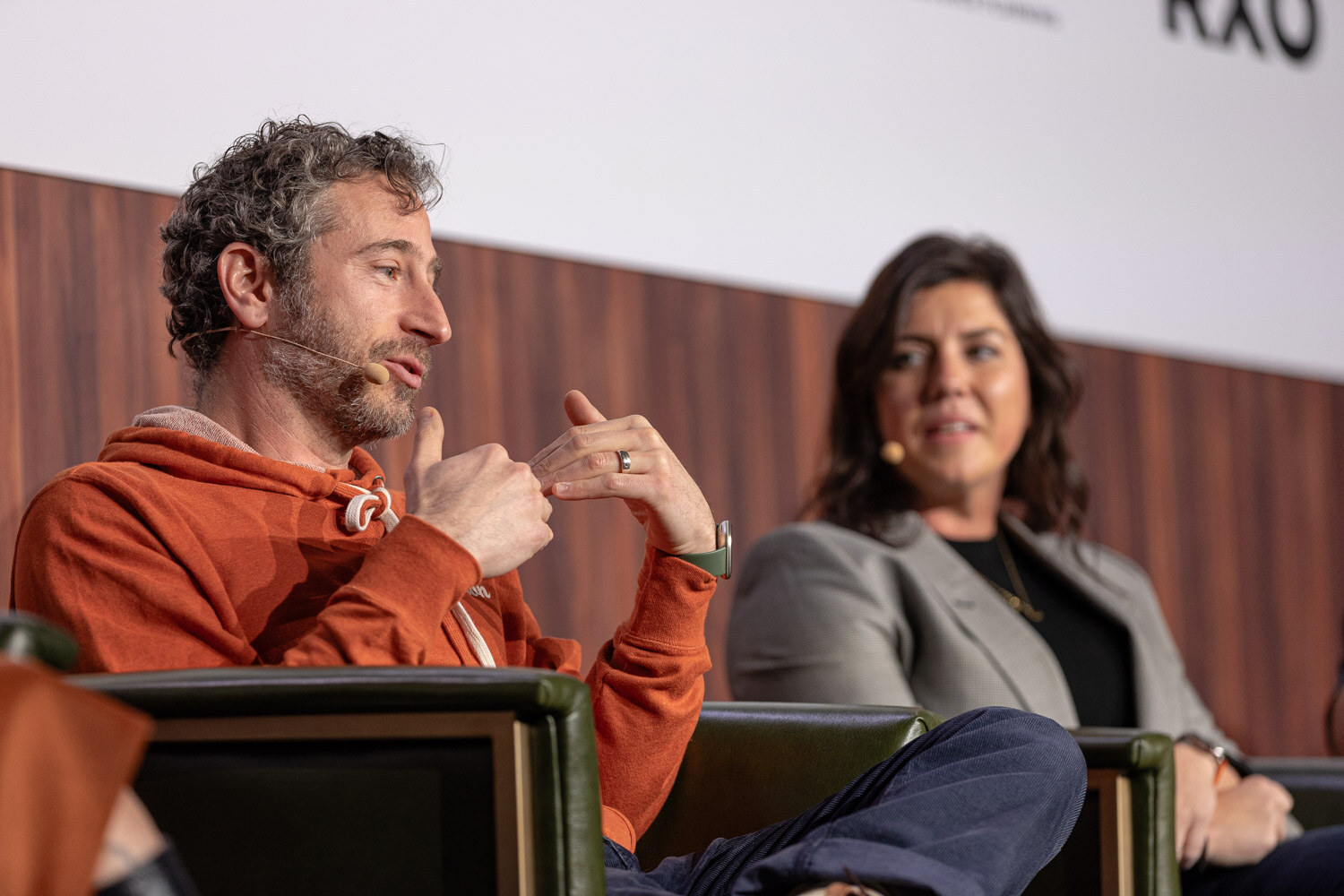 A curly-haired man in an orange hoodie gestures while a woman in a gray blazer sits beside him.