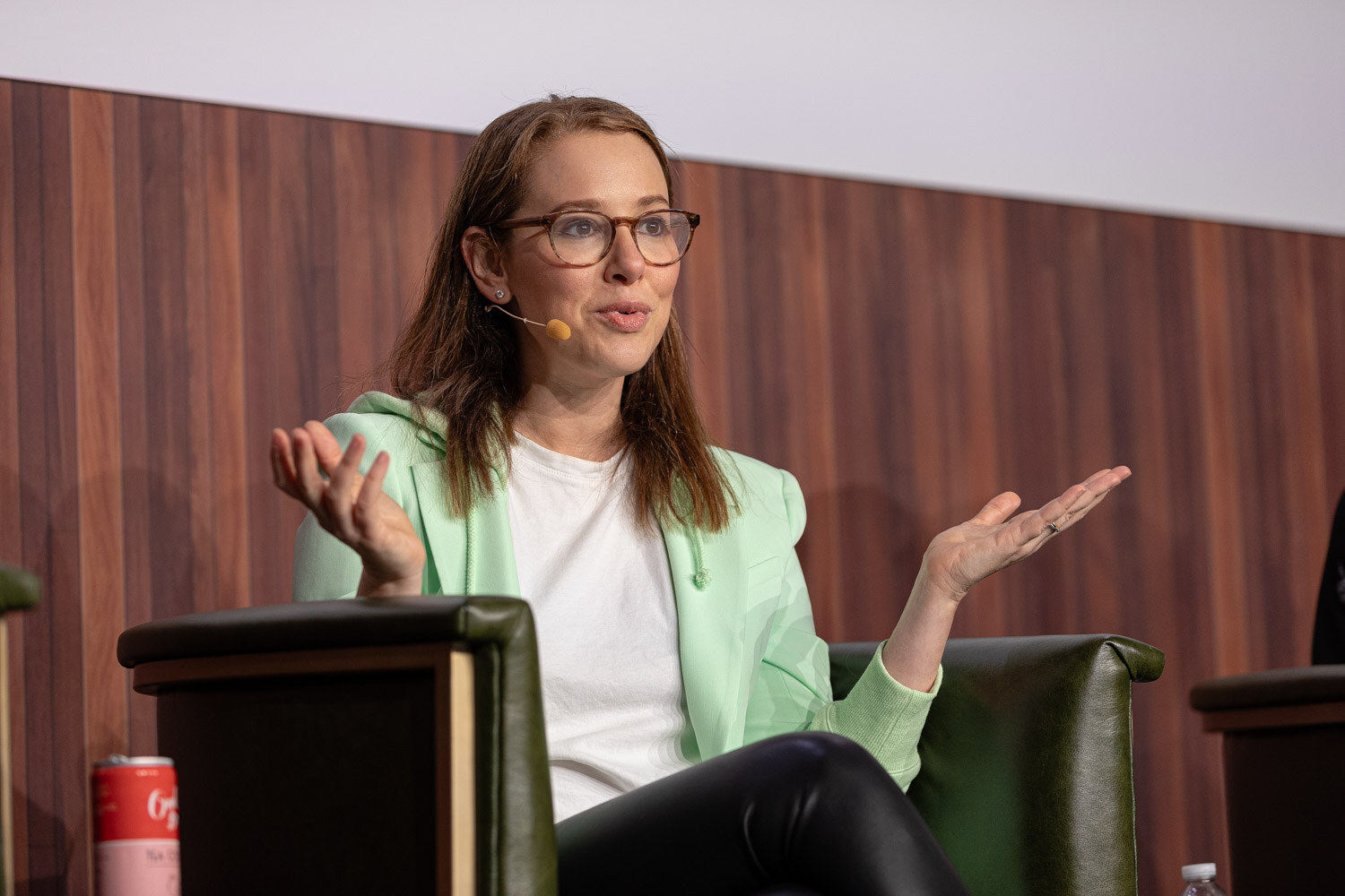 A woman with glasses and a mint green blazer gestures with open hands while seated in an armchair.