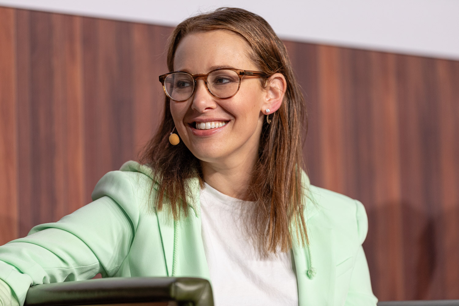 A smiling woman with glasses and a headset microphone wears a mint green blazer.