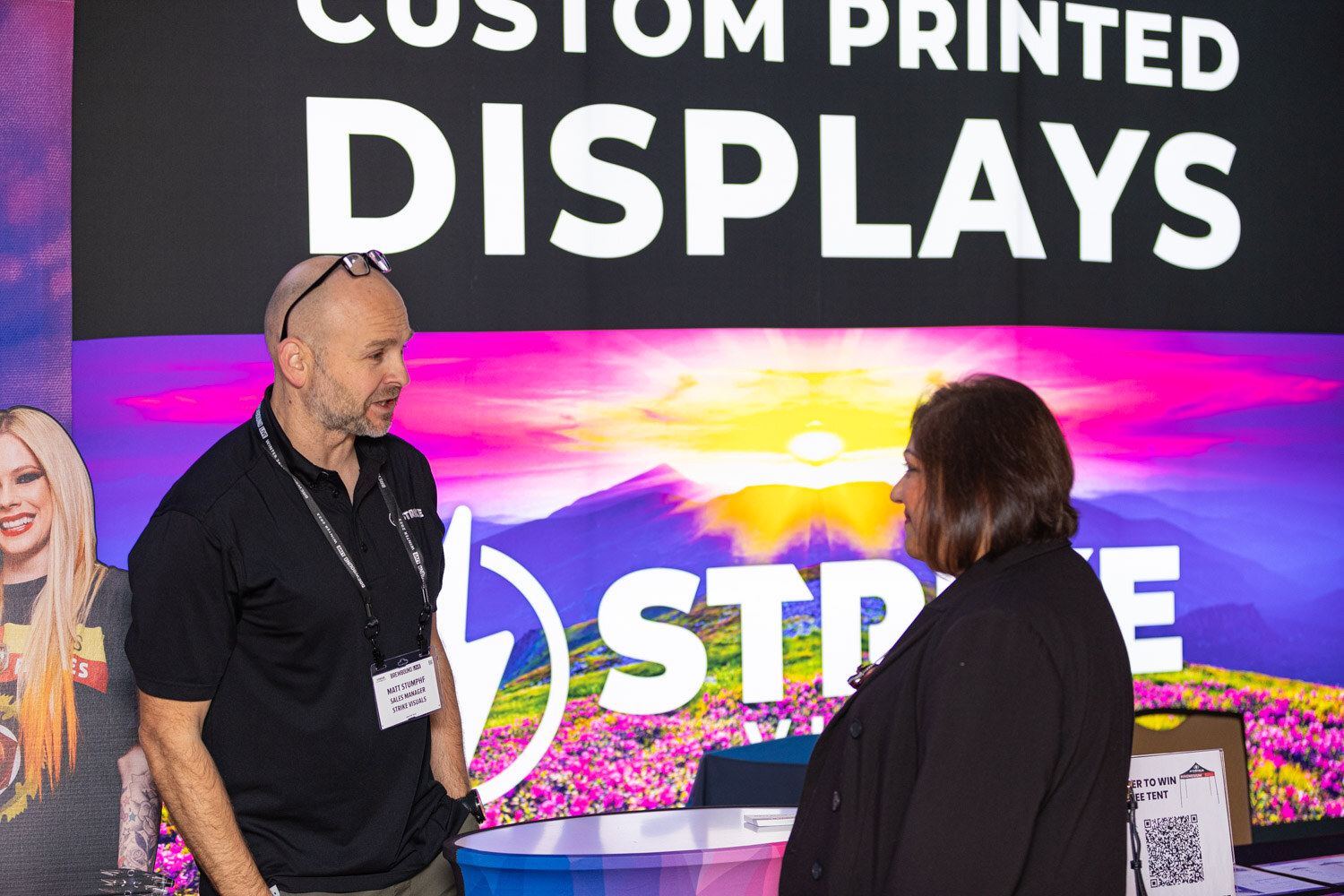 A bald man in a black polo with a lanyard talks with a woman in front of a Strike banner reading Custom Printed Displays.