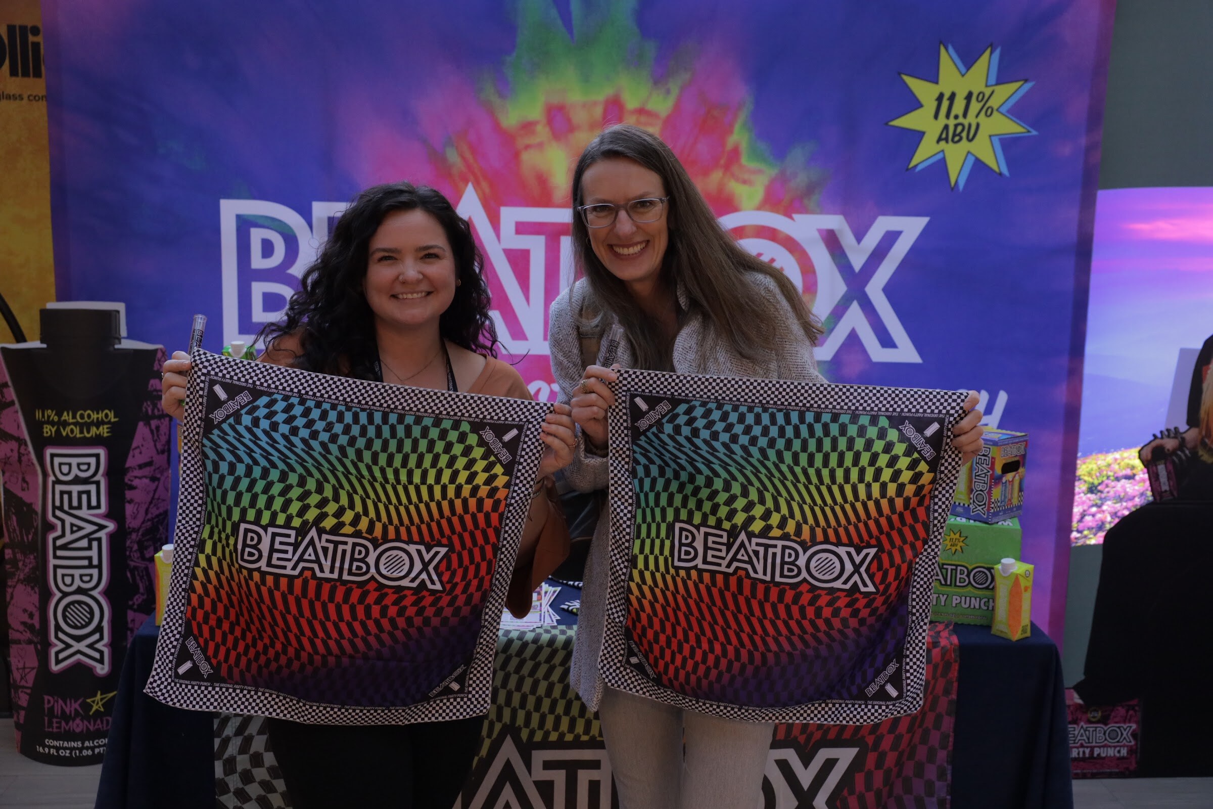 Two smiling women hold up rainbow BeatBox-branded bandanas in front of a BeatBox banner.