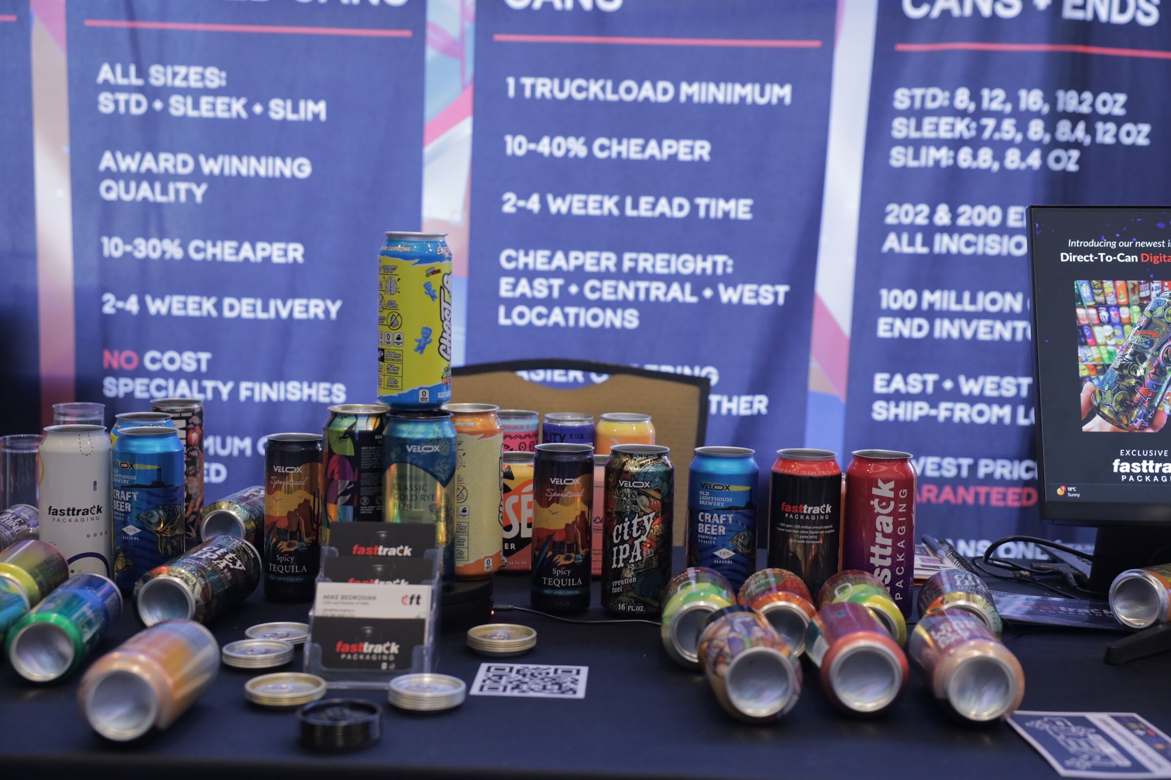 A table covered in colorfully printed aluminum cans in front of blue Fasttrack Packaging banners listing can sizes and lead times.