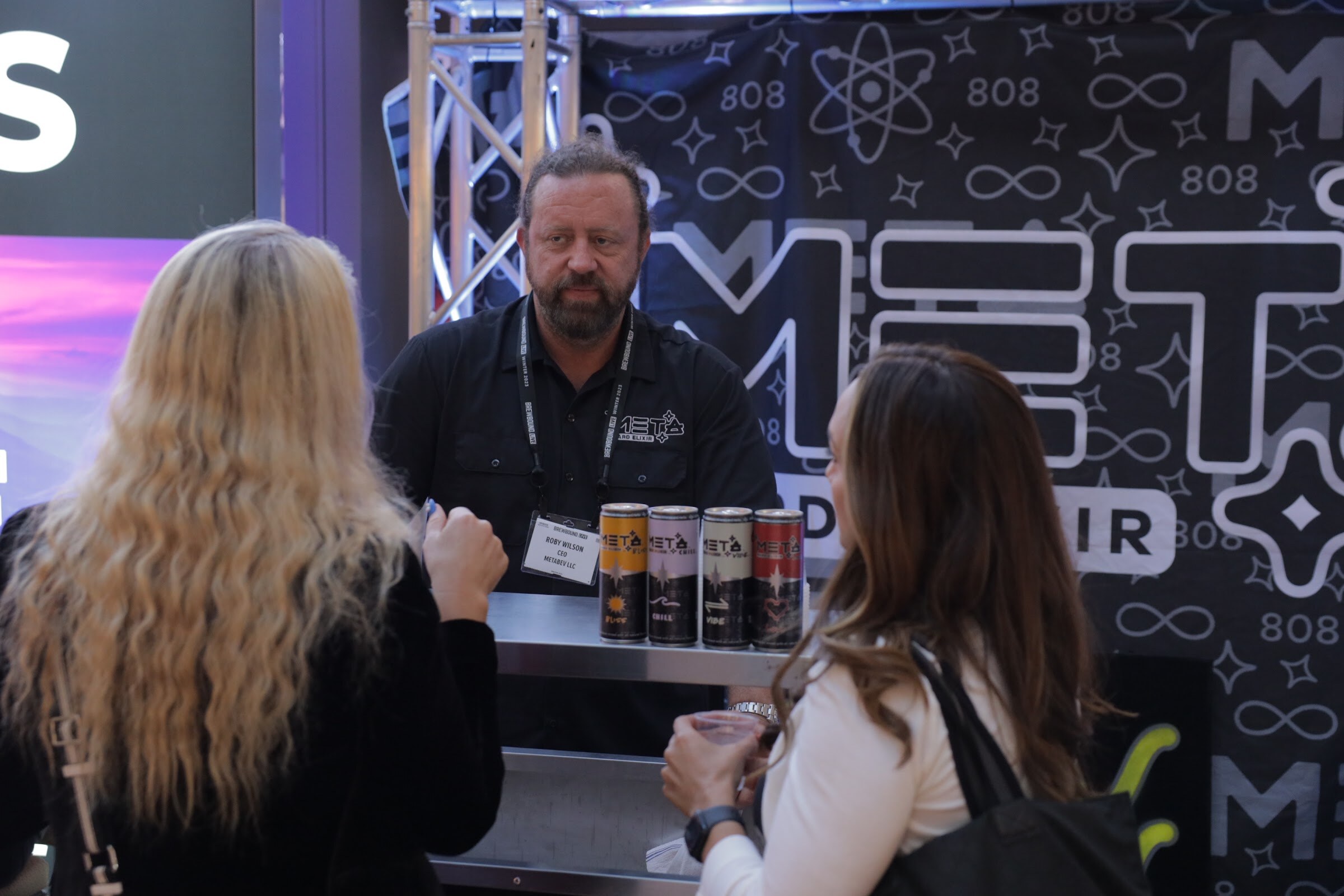 A bearded man in black stands behind a table of cans talking with two women in front of a black patterned backdrop.