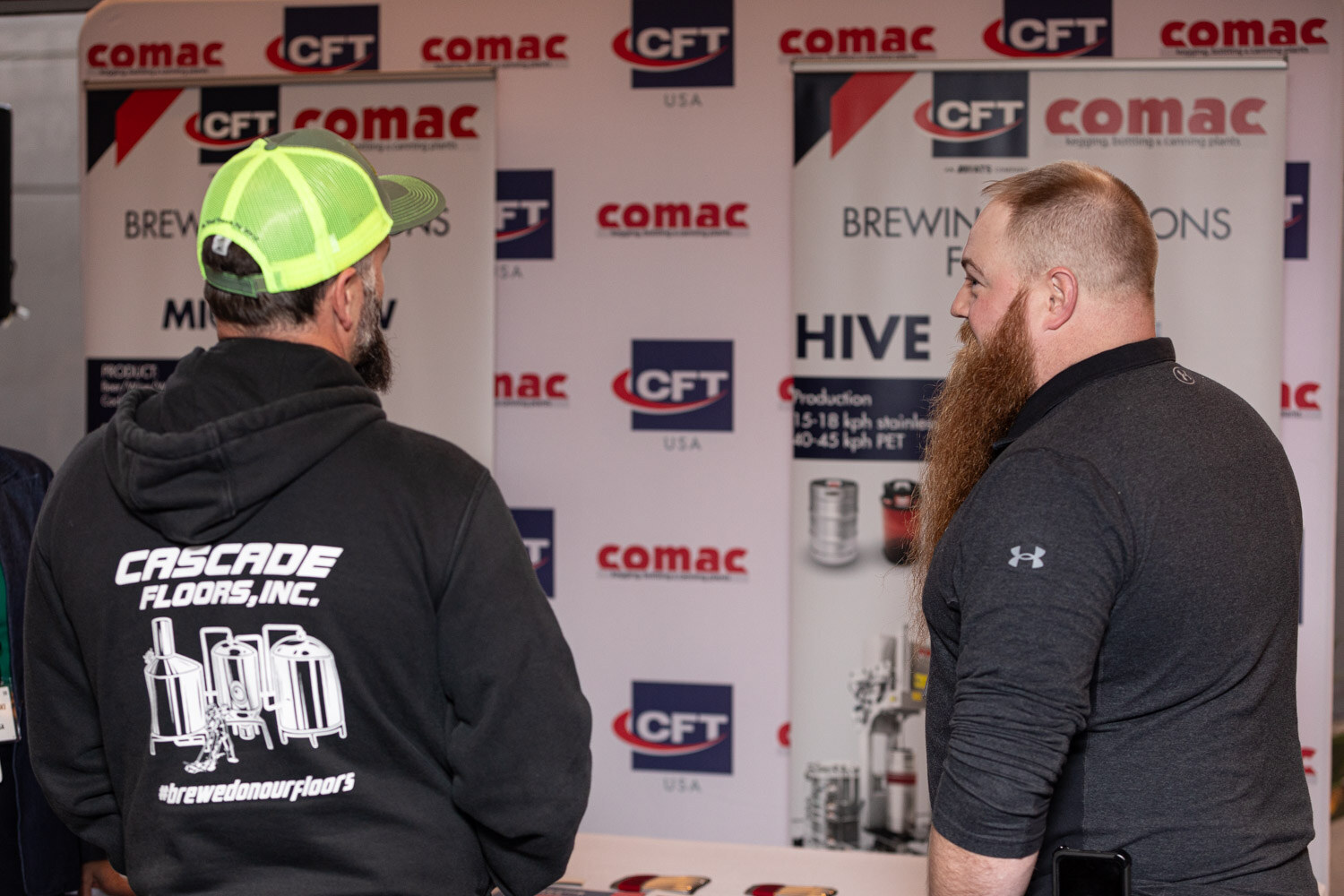 A man in a lime green cap and a black hoodie talks with a bearded man in a gray shirt in front of CFT and Comac banners.