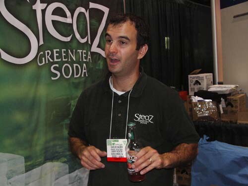 A man in a dark Steaz polo with a badge lanyard holds a bottle and speaks in front of a green Steaz Green Tea Soda banner. Boxes are stacked behind him.