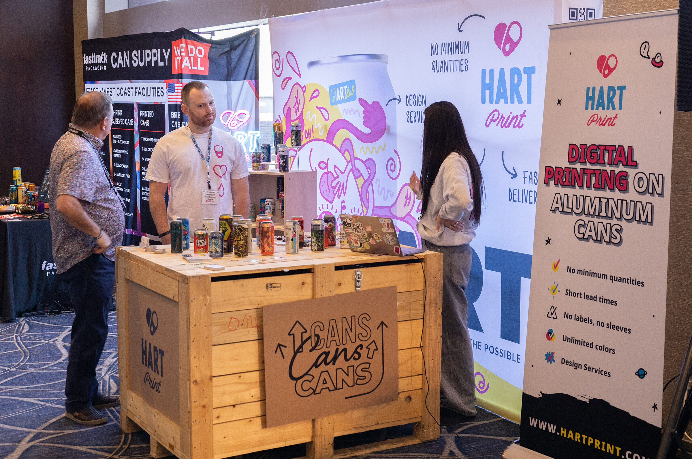 A wooden crate display with cans sits in front of Hart Print banners about digital printing on aluminum cans while people talk beside it.