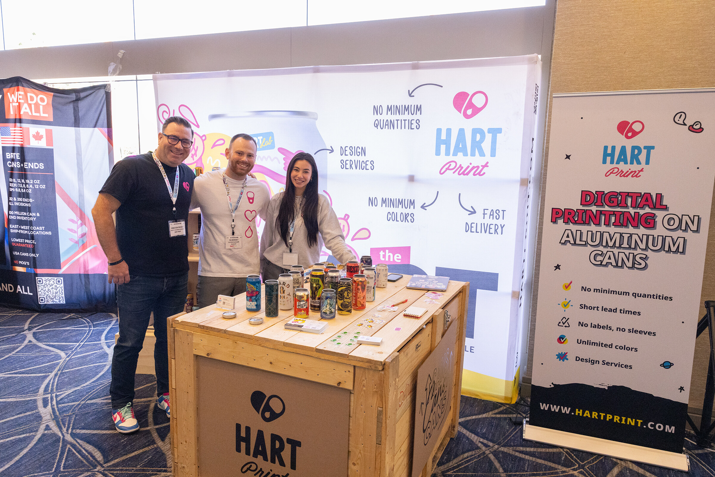 Three people smile at a wooden crate booth in front of Hart Print banners about digital printing on aluminum cans.