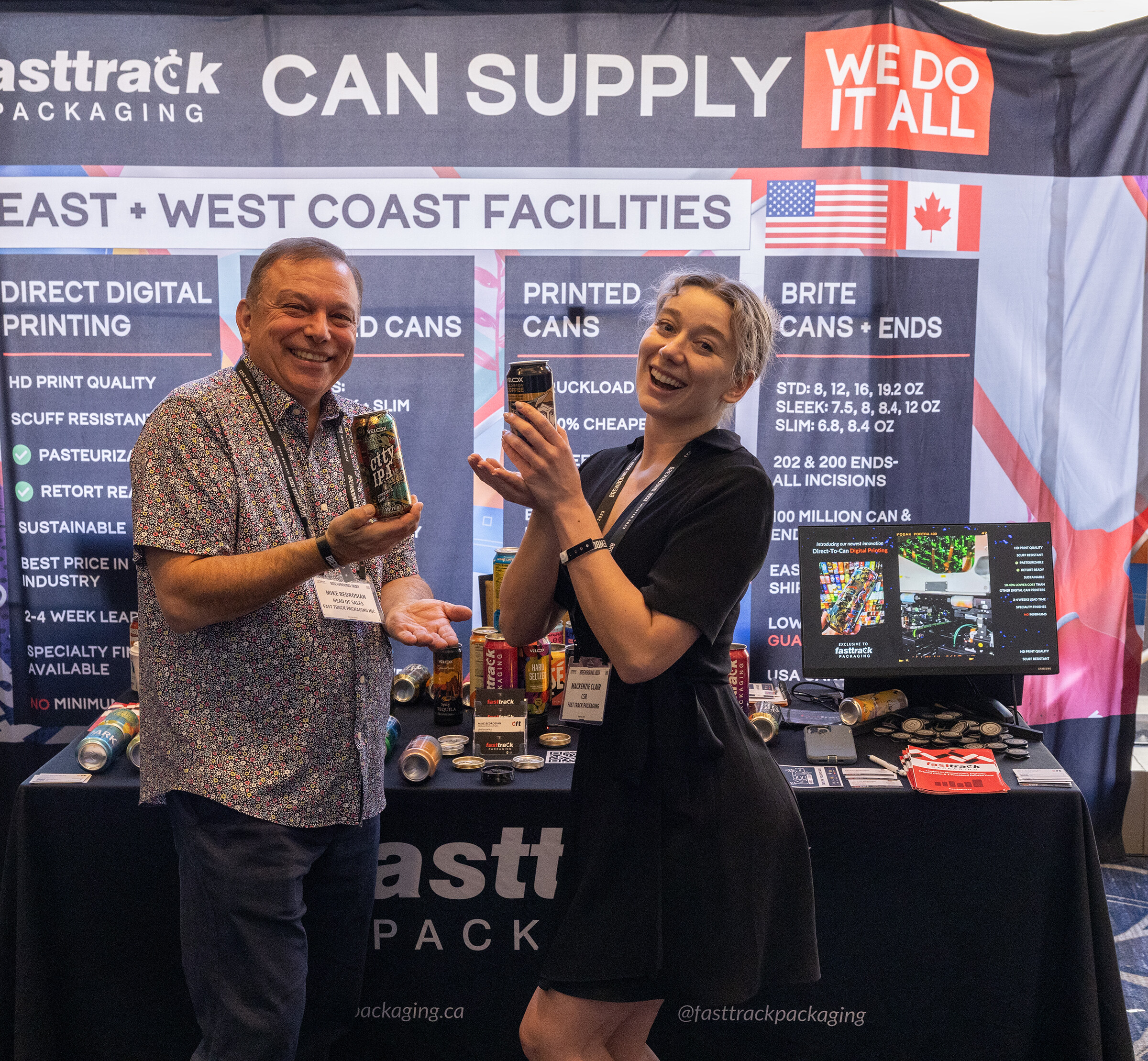 A smiling man in a patterned shirt and a laughing woman in black hold cans in front of a Fasttrack Packaging Can Supply backdrop.