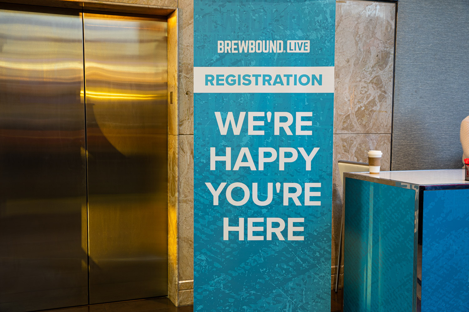 A teal Brewbound Live registration banner reading We're Happy You're Here beside brass elevator doors.
